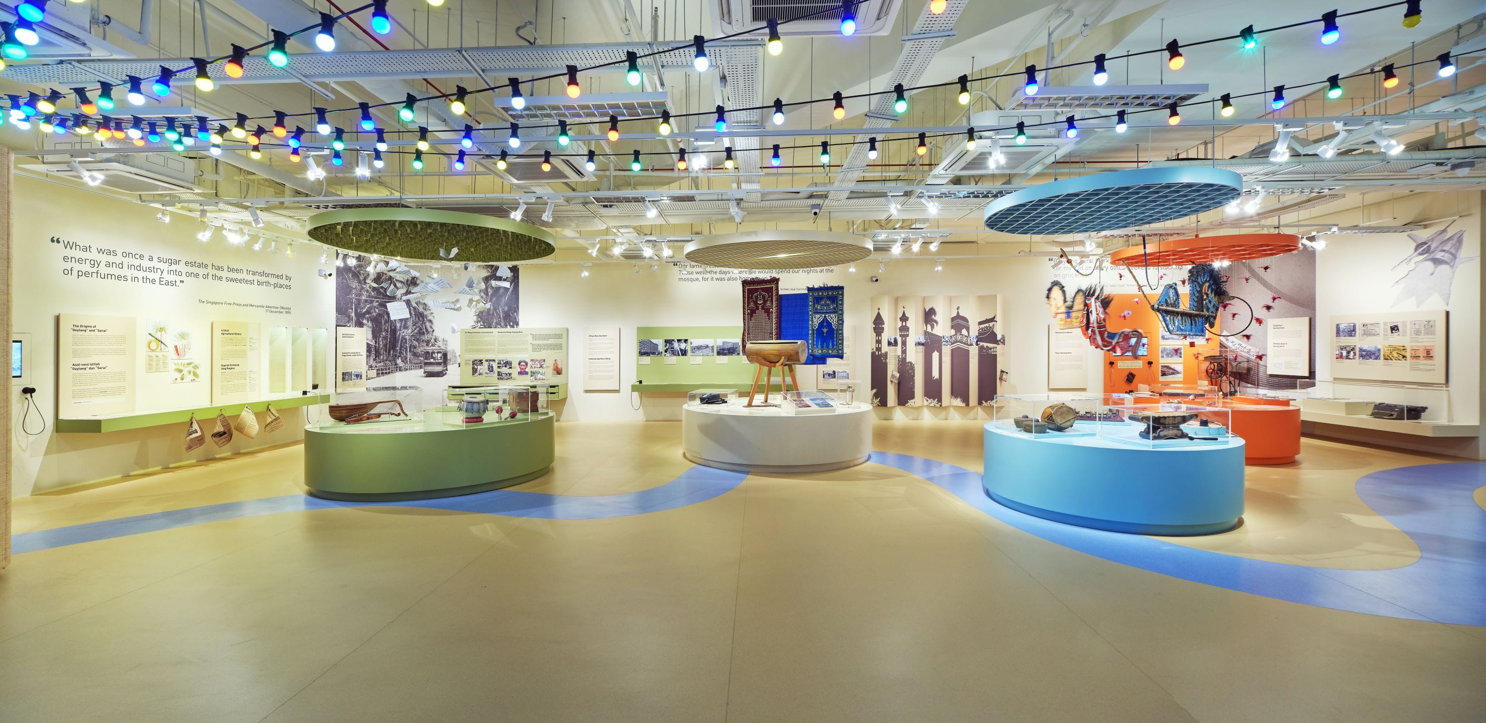 A colorful museum exhibit hall with round display cases, wall panels, hanging multicolored lights, and cultural artifacts on display.